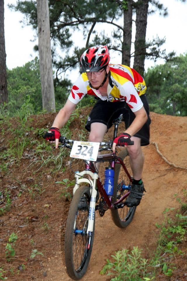 A cyclist in a colorful jersey and helmet rides a mountain bike down a dirt trail surrounded by trees. The bike has a number plate displaying "324," and the terrain is slightly hilly with green foliage visible. The cyclist appears focused and is navigating the trail skillfully. Flat Rock Park mountain bike trail.
