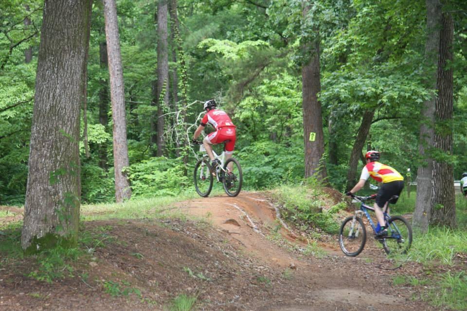 Two mountain bikers ride on a dirt trail through a lush green forest. One cyclist, wearing a red jersey, climbs a small incline, while the other, dressed in a yellow and black outfit, descends behind him. Tall trees and vibrant foliage surround the trail, creating a scenic outdoor environment. Flat Rock Park mountain bike trail.