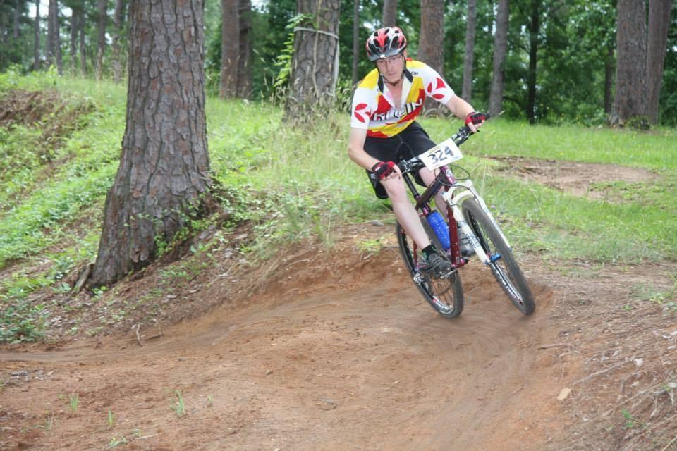 A person riding a mountain bike on a dirt trail, navigating a turn among trees and greenery. The cyclist is wearing a helmet and a colorful jersey with a race number visible on their shirt. Flat Rock Park mountain bike trail.