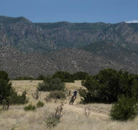 A person riding a mountain bike along a dirt trail in an arid landscape, with rugged mountains in the background and sparse vegetation surrounding the trail. Sandia Mountains Foothill Trail mountain bike trail.