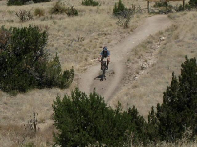 A person riding a mountain bike along a dirt trail in a grassy landscape, surrounded by bushes and sparse vegetation on a sunny day. Sandia Mountains Foothill Trail mountain bike trail.