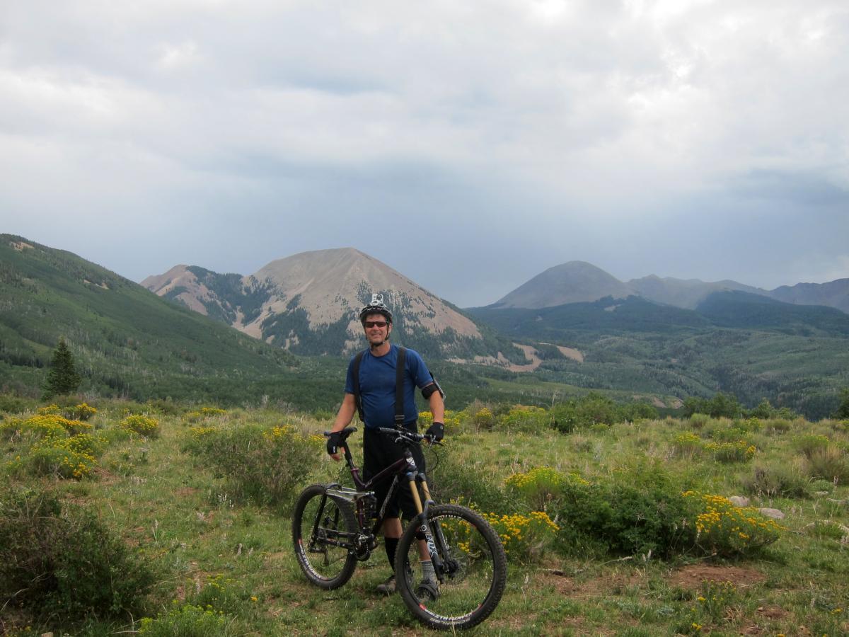 A mountain biker stands on a grassy hillside, smiling and holding his bike. The background features a panoramic view of lush green mountains under a cloudy sky, with patches of yellow wildflowers in the foreground. Hazard County mountain bike trail.