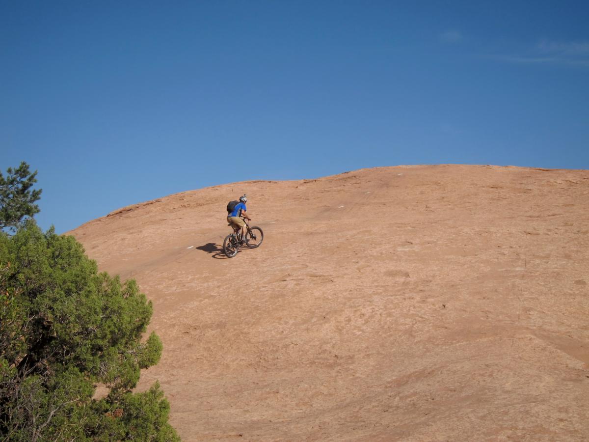 A mountain biker riding up a steep, sandy slope under a clear blue sky, with a patch of green vegetation in the foreground. Slickrock mountain bike trail.