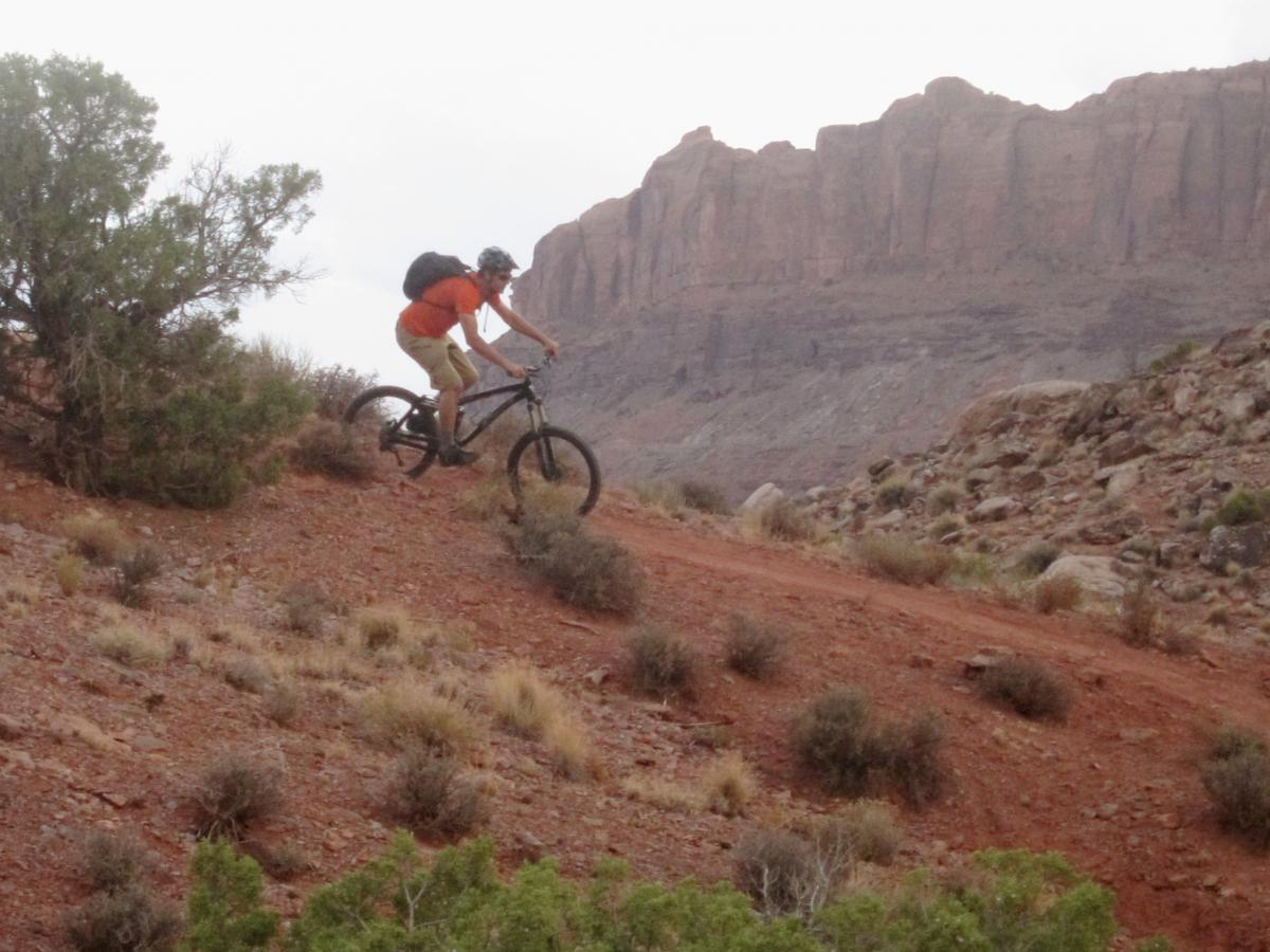 A mountain biker wearing an orange shirt and helmet rides along a dirt trail in a rugged landscape, surrounded by sparse vegetation and rocky terrain. Tall cliffs rise in the background under a cloudy sky. Moab Brand Trails mountain bike trail.