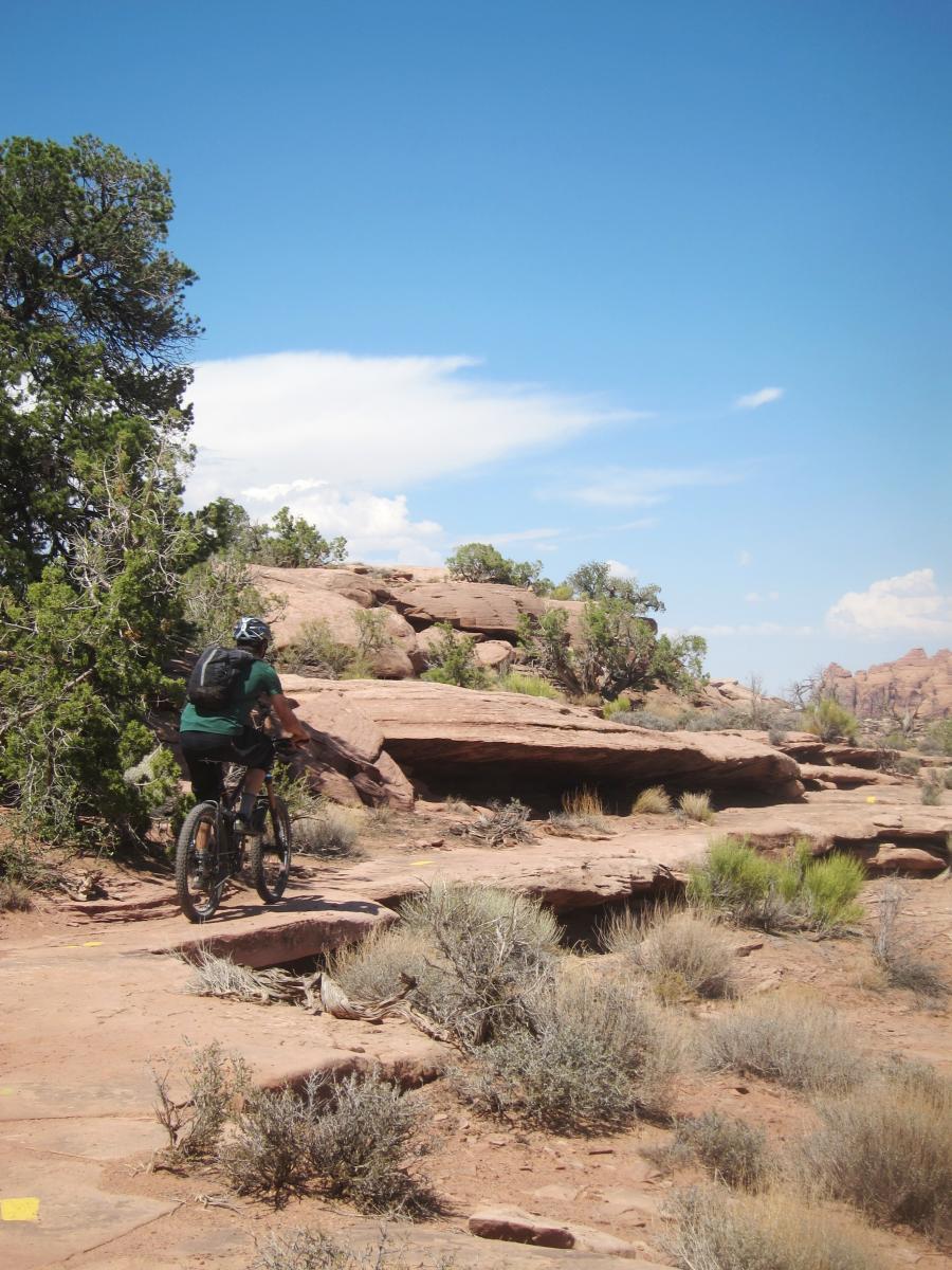 A person riding a mountain bike along a rocky trail surrounded by desert vegetation and large rock formations under a clear blue sky. Gemini Bridges mountain bike trail.