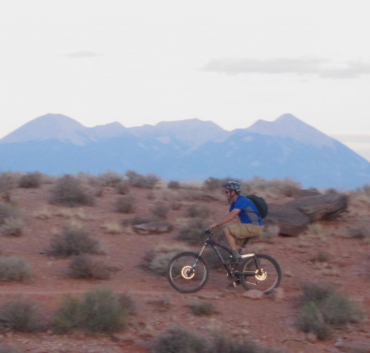 A mountain biker rides along a dirt trail in a desert landscape, with distant mountains in the background under a partly cloudy sky. The cyclist wears a blue shirt, shorts, and a helmet, carrying a backpack. Sparse vegetation and rocky terrain are visible around the trail. Moab Brand Trails mountain bike trail.