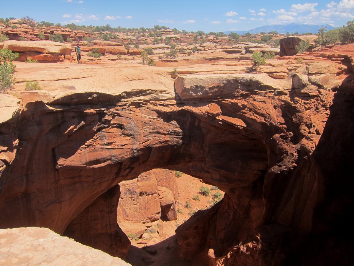 A hiker stands on a rocky ledge overlooking a natural stone arch in a desert landscape, with red sandstone formations and sparse vegetation under a blue sky with scattered clouds. Gemini Bridges mountain bike trail.