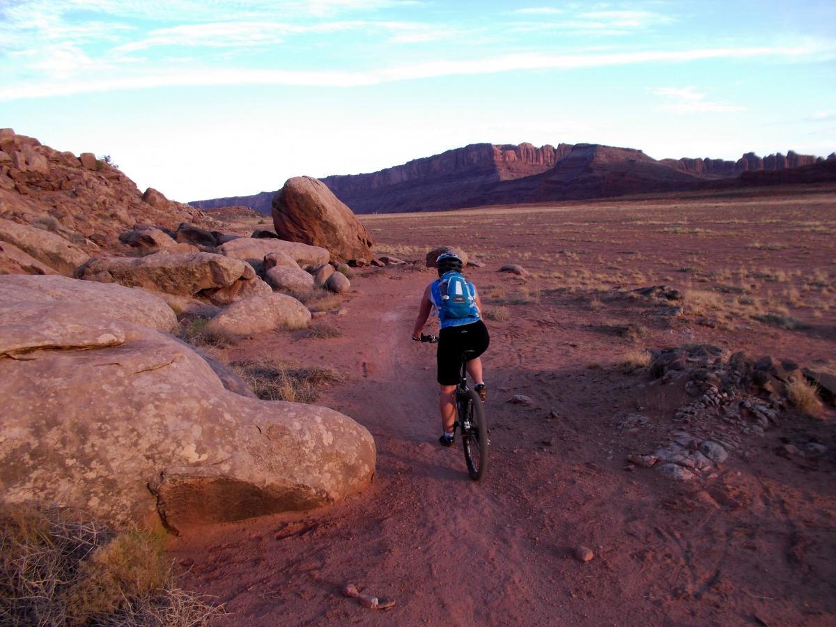 A cyclist rides along a dirt path surrounded by large rocks and a vast, arid landscape, with majestic cliffs in the background. The scene captures the beauty of nature during sunset, with warm tones illuminating the terrain. Moab Brand Trails mountain bike trail.