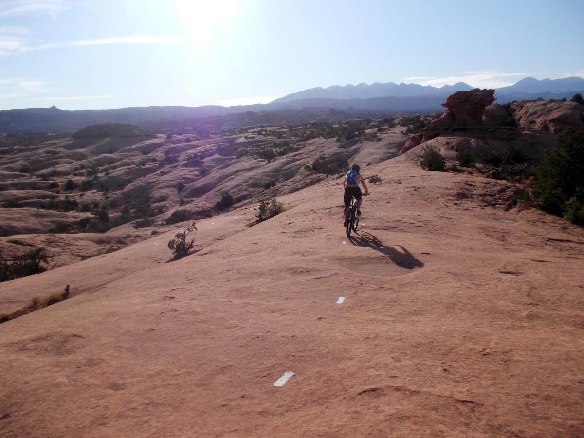 A mountain biker riding along a rocky terrain under bright sunlight, with distant mountains and blue skies in the background. The path features white markings guiding the way over the natural landscape. Slickrock mountain bike trail.