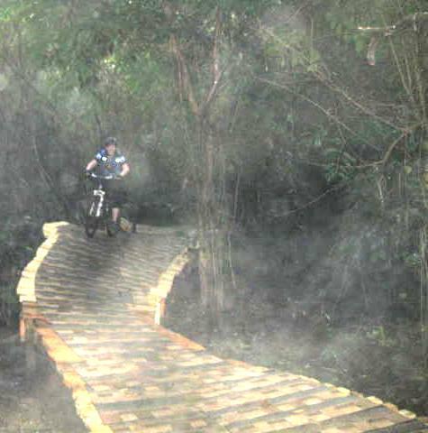A mountain biker riding a wooden trail through a misty, wooded area. The pathway is winding and elevated, surrounded by dense foliage. Markham Park mountain bike trail.