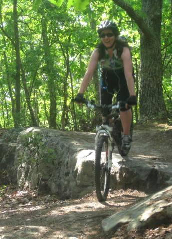 A person wearing a helmet and sunglasses is riding a mountain bike over a rocky trail surrounded by trees. The rider appears focused and is navigating the terrain with confidence on a sunny day. Raccoon Mountain Trail Network mountain bike trail.