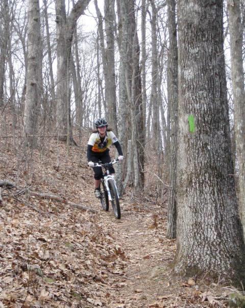 A mountain biker navigating a dirt trail through a wooded area, surrounded by bare trees and autumn leaves on the ground. A green marker can be seen on a tree trunk nearby, indicating the trail path. Aska Trail System mountain bike trail.