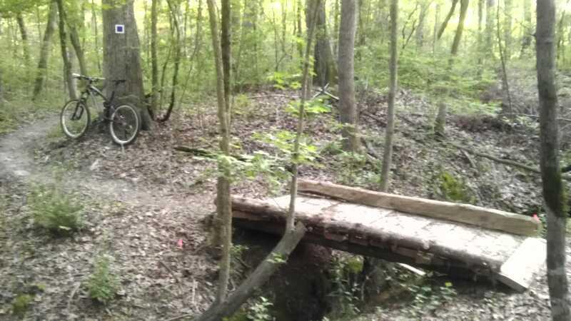 A mountain bike stands next to a dirt trail in a wooded area, with a rustic wooden bridge crossing over a small creek. The surrounding landscape features trees with green leaves and patches of sunlight filtering through the foliage. Small plants and fallen leaves cover the forest floor. River walk mountain bike trail.