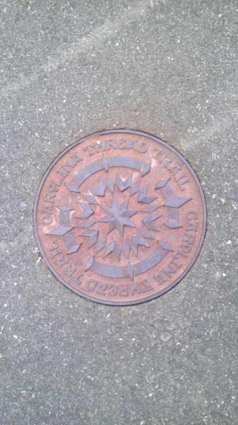 A circular metal manhole cover featuring a star design in the center, with engraved text around the edge that reads "Caution: Buried Hall." The cover is set on a textured concrete surface. River walk mountain bike trail.