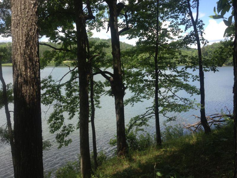 A serene view of a lake surrounded by trees, with sunlight filtering through the leaves. The calm water reflects the blue sky and distant hills, creating a peaceful natural setting. Issaqueena Lake mountain bike trail.