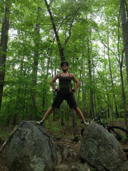 A person stands triumphantly on two large rocks in a forested area, surrounded by green trees. They are wearing a helmet and a sleeveless top, with their hands on their hips and legs spread apart, exuding a sense of accomplishment. A mountain bike can be seen nearby, partially obscured by the vegetation. San-lee Park mountain bike trail.