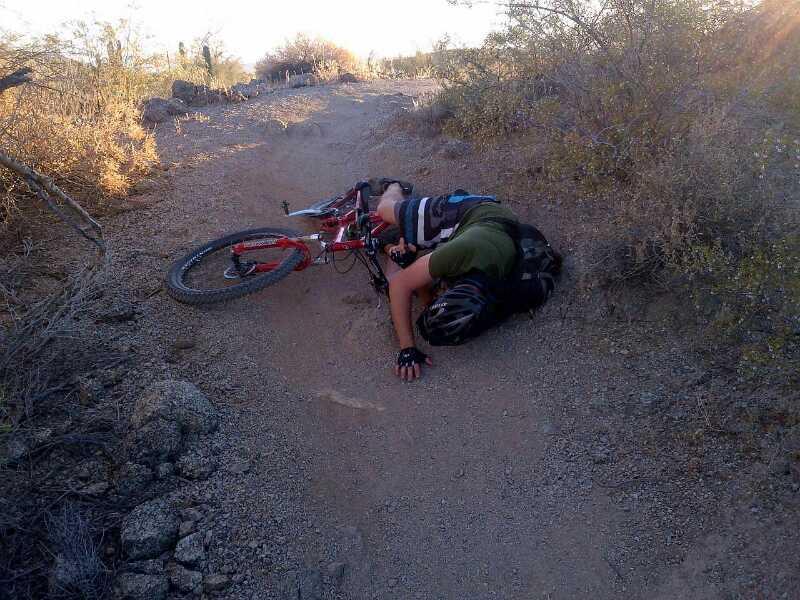 A mountain biker lying on the ground beside a fallen bicycle on a dirt trail, surrounded by sparse vegetation and rocky terrain, indicating a potential fall or accident. Hawes Loop mountain bike trail.