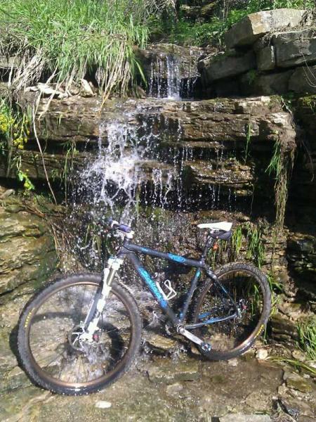 A mountain bike positioned near a small waterfall cascading over rocky terrain, surrounded by lush green grass and plants. Water splashes around the bike, highlighting a natural outdoor setting. Decorah Mtb Trail System mountain bike trail.