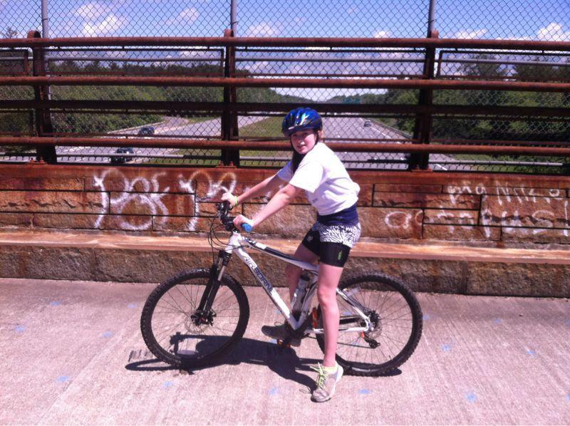 A young person wearing a white T-shirt and a blue helmet is sitting on a mountain bike on a bridge. In the background, there is a clear blue sky with a few clouds and greenery. The bridge has a rusty railing with graffiti on the wall beside it. A roadway is visible below the bridge. Georgetown-rowley State Forest mountain bike trail.