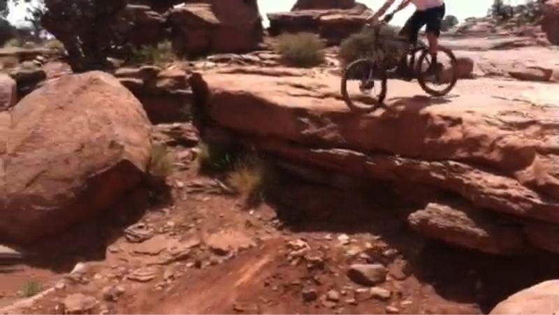A mountain biker navigating a rocky terrain, with large boulders and rugged ground in the surrounding landscape. The biker is positioned on a ledge, showcasing an action moment of the ride. Captain Ahab mountain bike trail.