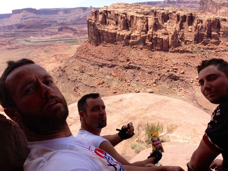Three men taking a selfie while sitting on a rocky ledge, overlooking a vast canyon landscape with large rock formations in the background. The sky is clear with some clouds, creating a sunny and open atmosphere. One of the men is holding a snack. Captain Ahab mountain bike trail.