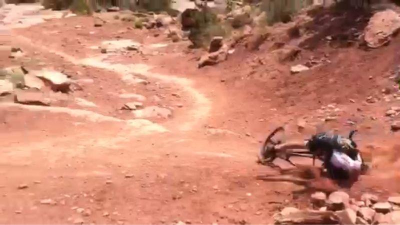 A person falls off a mountain bike while riding on a rocky dirt trail, surrounded by sparse vegetation and boulders. The trail curves in the background. Captain Ahab mountain bike trail.