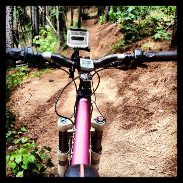 Close-up view of a mountain bike's handlebars, featuring a digital display and a camera mounted on top. The trail ahead winds through a densely wooded area with green foliage and pine needles on the ground. Forks Area Trail System (FATS) mountain bike trail.