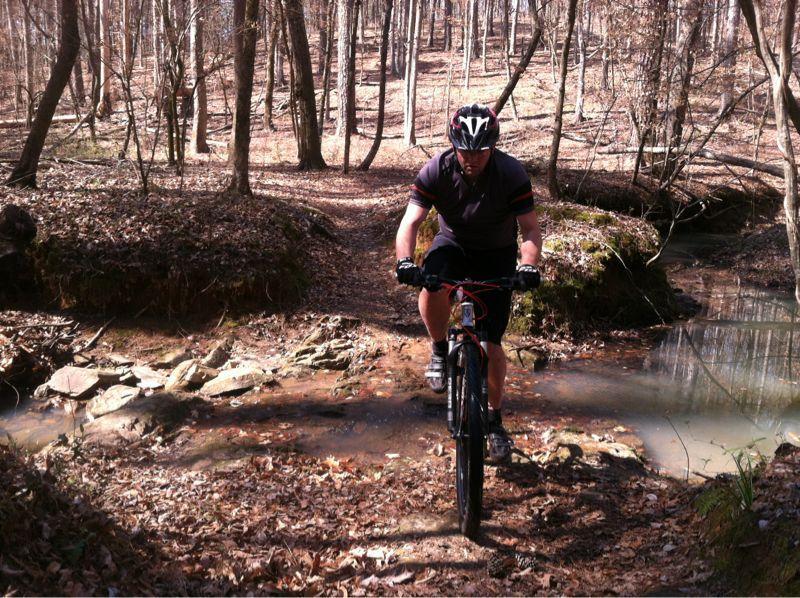 A mountain biker navigating through a rocky area near a small stream in a wooded forest. The trail is covered with fallen leaves and sunlight filters through the trees, creating a natural outdoor setting. The biker is wearing a helmet and cycling gear, focused on maintaining balance as he rides over the uneven terrain. Modoc (Stevens Creek Trail) mountain bike trail.
