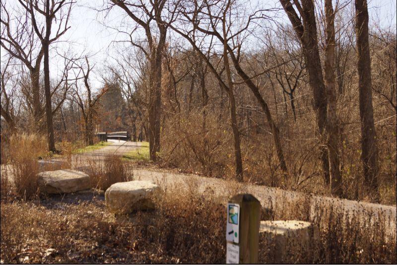 A pathway through a wooded area, featuring bare trees and dried vegetation. In the background, there's a wooden footbridge crossing over a small creek, with a sign on the right indicating a trail. Sunlight filters through the branches, creating a serene outdoor setting. Al Foster mountain bike trail.
