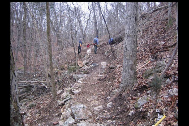 A group of people working on a rocky trail in a wooded area, surrounded by bare trees and fallen leaves. Some individuals are tending to the trail, while tools and equipment are scattered nearby. The scene conveys a sense of outdoor maintenance and teamwork. Lost Valley mountain bike trail.