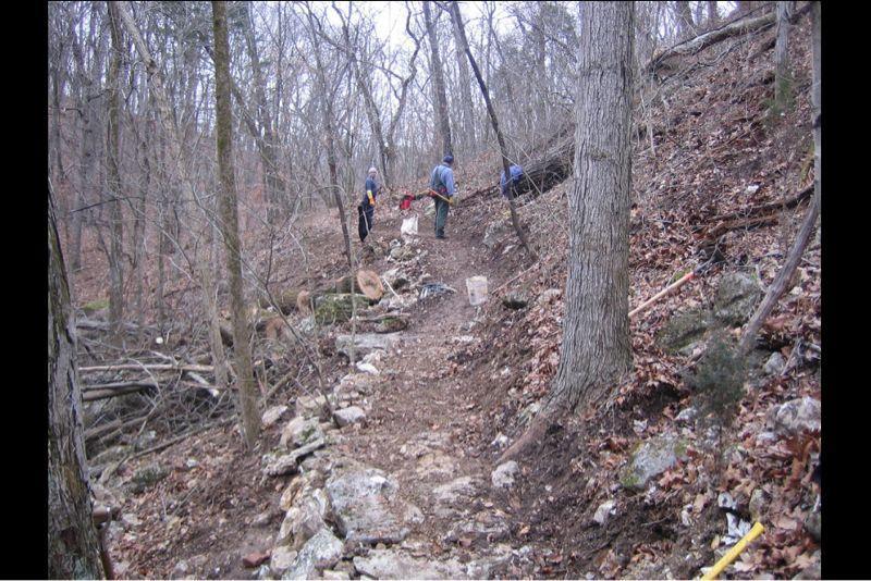 A rocky hiking trail winding through a wooded area with several people working in the background, surrounded by bare trees and fallen leaves. Lost Valley mountain bike trail.