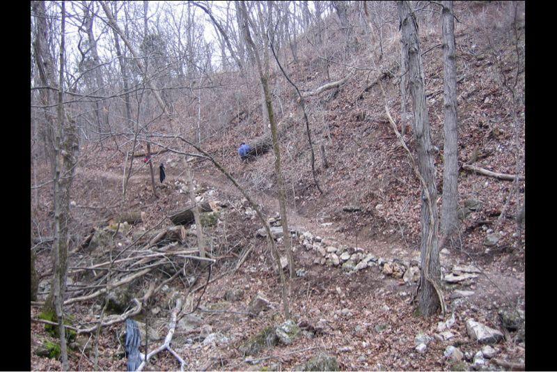 A wooded area with a hillside covered in bare trees and scattered fallen branches. A narrow dirt trail winds through the landscape, with a stone path leading along the trail. In the background, a person in a blue shirt is seen near the base of the hill, engaging in some task amidst the natural surroundings. The scene appears to be in late fall or early spring, with no foliage on the trees. Lost Valley mountain bike trail.