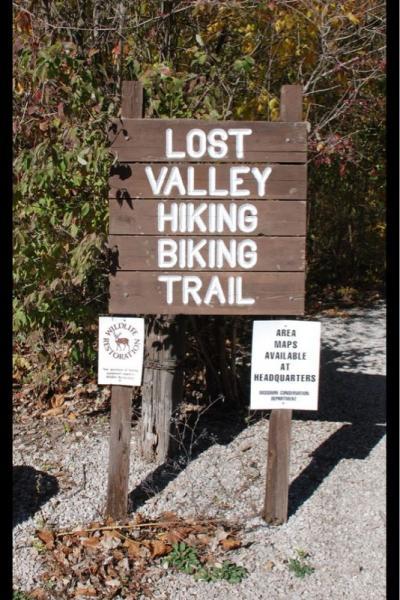Wooden sign at the entrance of the Lost Valley Hiking and Biking Trail, surrounded by trees and shrubs. The sign features the trail name prominently, along with additional information about area maps being available at headquarters. Lost Valley mountain bike trail.