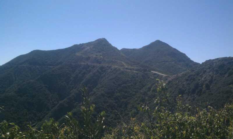A scenic view of two mountainous peaks surrounded by lush greenery under a clear blue sky. A winding road can be seen traversing the slopes of the mountains. Romero Road mountain bike trail.
