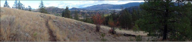 A panoramic view of a hillside with tall grass and scattered trees, overlooking a valley with a small town in the distance. Rolling hills and mountains are visible under a partly cloudy sky. A narrow dirt path winds through the grass in the foreground. MT Conkle mountain bike trail.