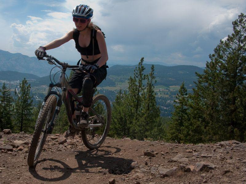 A person riding a mountain bike on a rugged trail with a scenic view of mountains and trees in the background. The cyclist is wearing a helmet and protective gear, and the sky is partly cloudy. Smith Creek mountain bike trail.
