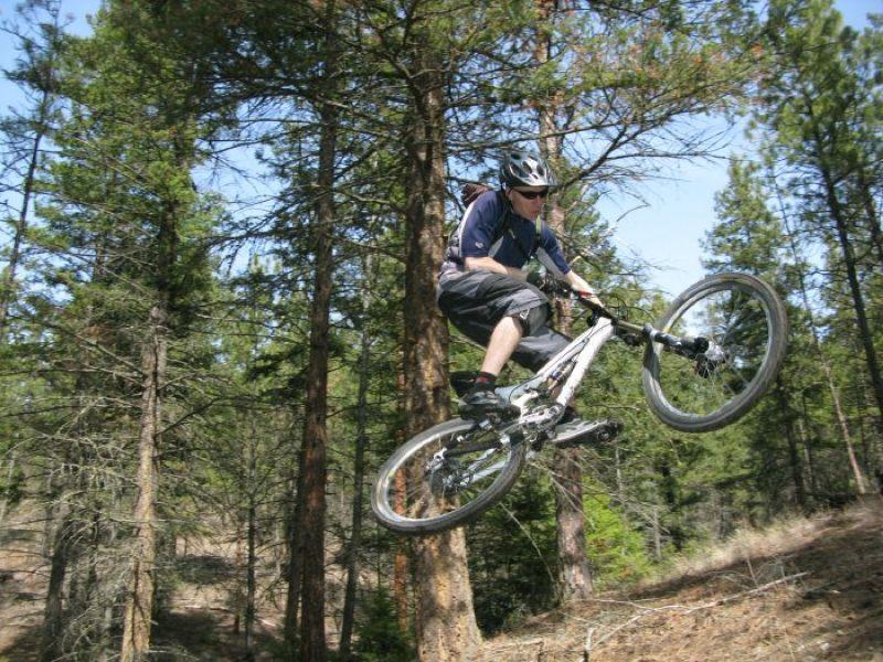 A mountain biker in mid-air performing a jump on a forest trail, surrounded by tall pine trees under a clear blue sky. The rider is wearing a helmet and protective gear, showcasing an adventurous outdoor activity. Smith Creek mountain bike trail.