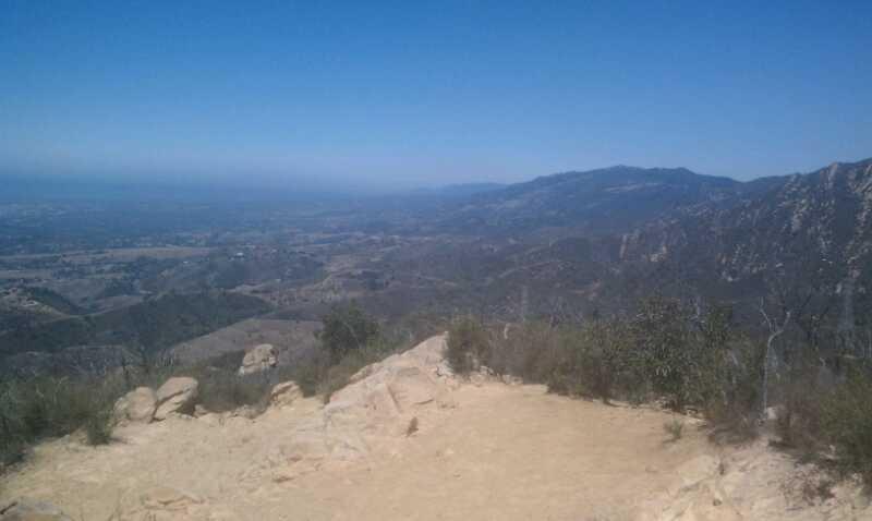 A panoramic view from a mountain overlook, showcasing rolling hills and valleys under a clear blue sky. The foreground features rocky terrain with sparse vegetation, while distant mountains create a scenic backdrop. Jesusita mountain bike trail.