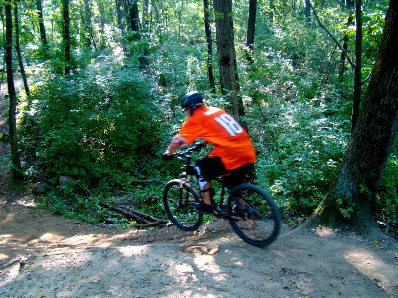 A person wearing an orange jersey with the number 18 rides a mountain bike along a dirt trail in a forested area, surrounded by greenery and trees. Palos Forest Preserve mountain bike trail.