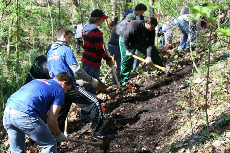 A group of people working together in a wooded area, using tools to dig and clear a trail. The scene captures teamwork and community involvement in outdoor activities, with individuals dressed in casual clothing and surrounded by greenery. Bluffview trail mountain bike trail.