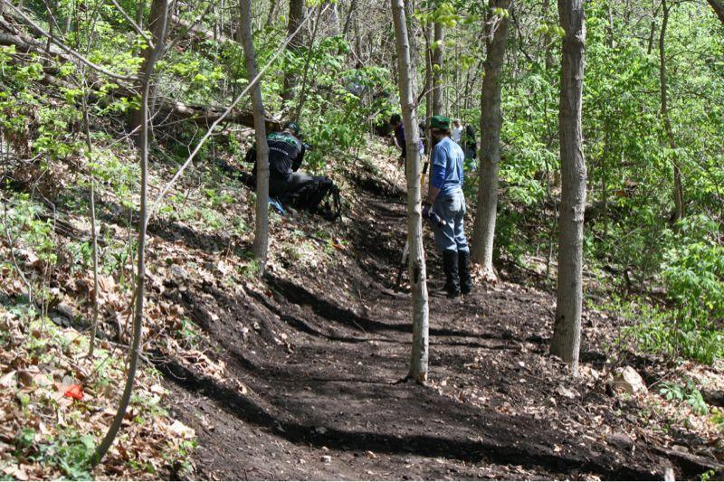A group of individuals working on a trail in a wooded area, surrounded by trees and greenery. The path appears to be recently cleared, with dirt showing along the trail. Some people are engaged in labor, while others are observing the work. Bluffview trail mountain bike trail.