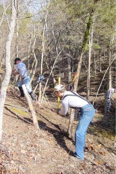 Two men working in a wooded area, using tools to clear or manage the landscape. One man is chopping at a tree stump with an axe, while the other is holding a tool and crouched down near another stump. The scene depicts an outdoor environment with sparse trees and fallen leaves on the ground. Chubb Trail mountain bike trail.