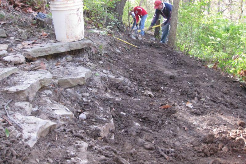 Two people working on landscaping in a wooded area. The foreground shows a set of stone steps and a patch of disturbed earth, while the background features trees and greenery. One person is using a shovel, while the other is measuring with a tape. A bucket is placed nearby among the rocks and soil. Chubb Trail mountain bike trail.