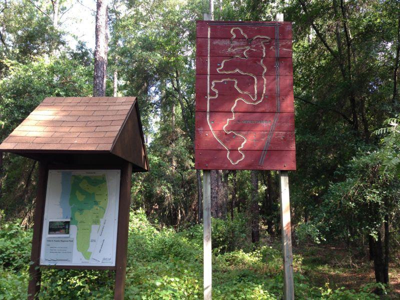 A wooden signpost displaying a map of a nature park is situated among lush greenery. To the left, a smaller map board provides information about the park's layout, while a larger red sign on the right features a stylized representation of the park's trails or features. The scene is surrounded by trees and dense foliage, indicating a natural outdoor setting. Tillie Fowler Regional Park mountain bike trail.