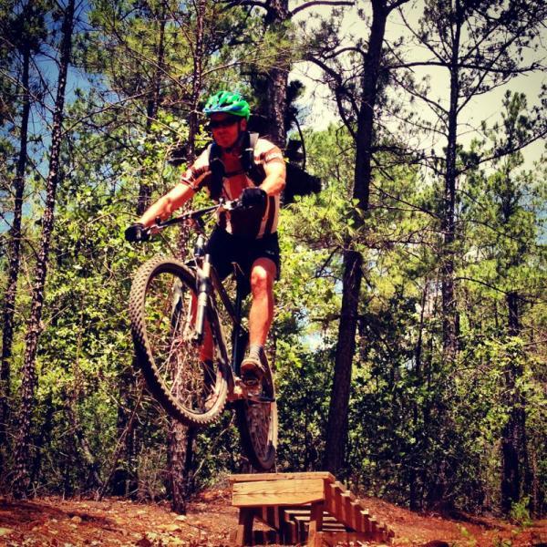 A mountain biker in a green helmet is mid-jump on a trail feature, surrounded by trees. The rider is wearing a light-colored shirt and shorts, and a backpack is mounted on their back. The background shows a mix of green foliage and blue sky. Rocky Hill Ranch mountain bike trail.