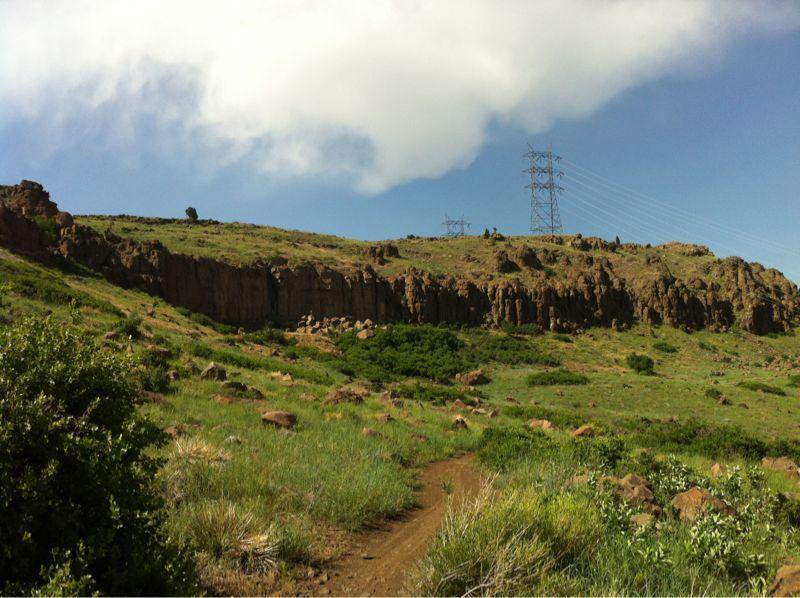 A scenic view of a green hillside with rocky cliffs under a partly cloudy sky. Power lines are visible in the background, and a dirt path winds through the grassy landscape. North Table Mountain mountain bike trail.