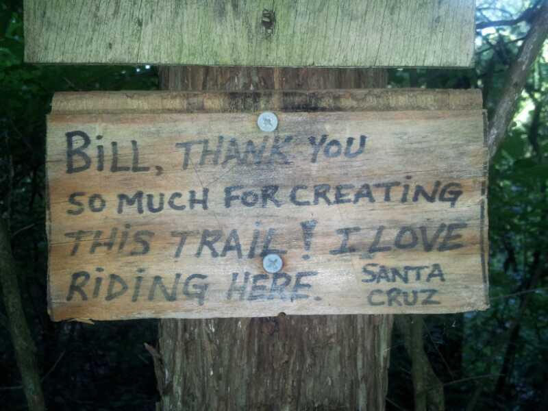 Wooden sign attached to a tree, expressing gratitude to someone named Bill for creating a trail. The message reads: "Bill, thank you so much for creating this trail! I love riding here. Santa Cruz." The sign has visible screws and is surrounded by greenery. Milford Trail mountain bike trail.