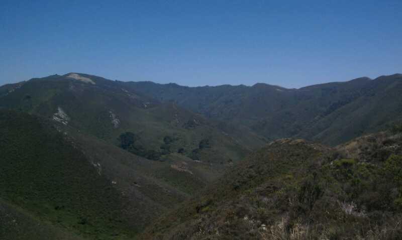 A panoramic view of rolling green hills and mountains under a clear blue sky, showcasing a lush, natural landscape with varying shades of green and sparse vegetation in the foreground. Montana De Oro mountain bike trail.