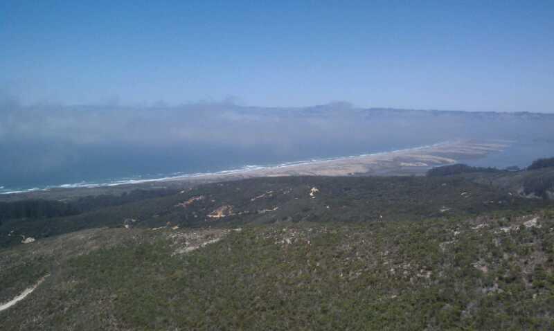 A panoramic view overlooking a coastal landscape with rolling hills, dense greenery, and a sandy beach meeting the ocean's waves. The sky is clear with a few scattered clouds, showcasing the vastness of the sea and the distant coastal horizon. Montana De Oro mountain bike trail.