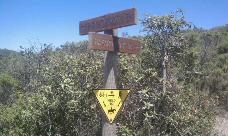 Signpost at a hiking trail junction indicating directions for East Bolinas Trail and Hazard Rock Trail, accompanied by a yellow yield sign instructing users to yield to cyclists and horseback riders. Surrounded by lush greenery and clear blue skies. Montana De Oro mountain bike trail.
